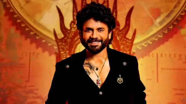 The host of "Bigg Boss Telugu" stands confidently against a themed backdrop.